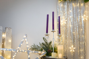 Christmas decorations. Purple candles in candlestick, fir tree branches and cones, siver curtains with shiny stars and warm garland lights and bokeh.