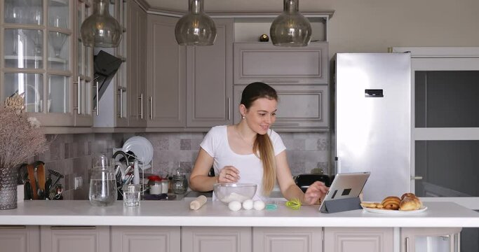 young woman cooking dough for pancakes or cookies and watching online recipe on a tablet in the kitchen.