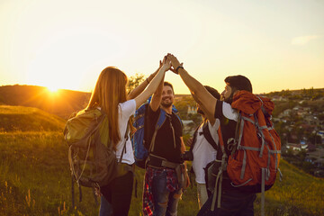 Group of hikers.Team of happy tourists with backpacks raised their hands up in nature at sunset in...