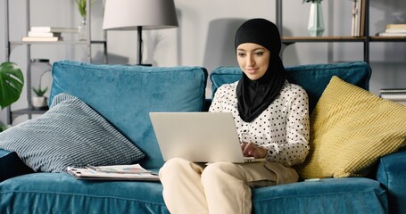 Portrait of cheerful muslim young woman at home, working remotely while sitting on couch in living room with laptop computer.