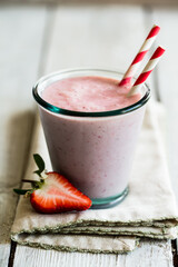 Vegan friendly strawberry and oat milk smoothie served in a green recycled glass with paper straws and fresh strawberry on a napkin on white wooden worktop