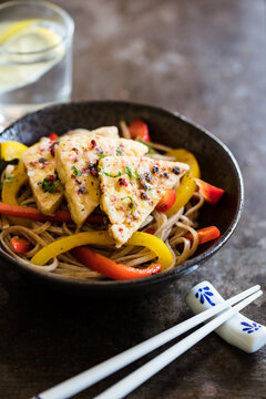 Salt and pepper tofu on a bed of  stir fried vegetables and noodles served in a bowl with chopsticks on rustic background