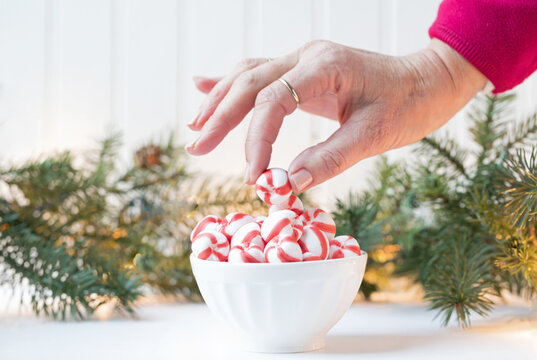 Original Christmas Photograph Of A Woman’s Hand Picking Up A Red And White Peppermint Candy Out Of A White Bowl With Christmas Greens And Lights