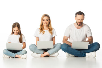 cheerful family sitting on floor with laptops on crossed legs isolated on white