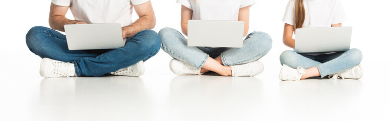 cropped view of family sitting on floor with laptops on crossed legs on white, panoramic shot