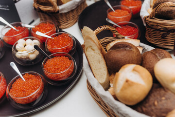 Bowls with red caviar and balls of butter located on black trays on a table served from a basket with different types of bread.