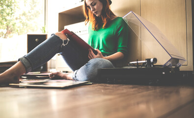 A woman sitting on the floor next to a record player looking at a record sleeve.