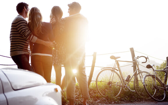Four people standing by the side of a road, parked car and bicycles leaning against a fence, sunlight.