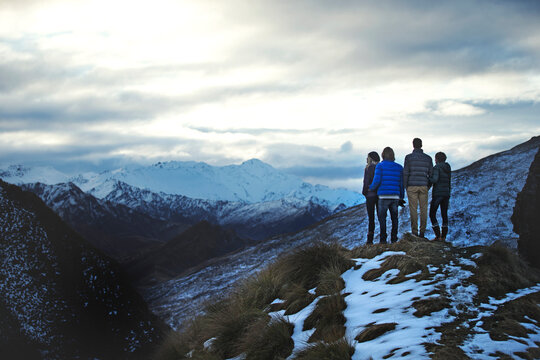 Rear view of four people standing side by side on a mountain, snow-capped peaks in the distance.