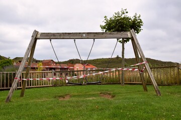 Fototapeta premium Swings in a playground in Spain closed by the Sars-Cov-2 pandemic (covid19). Its use is prohibited by placing plastic tape. In the background houses from the village of Clavijo.