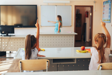 Two little schoolgirls sit at a desk in a school class and raise their hands