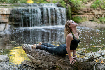 Blonde woman in sportswear practicing yoga on a log at the forest riverside in front of a waterfall. Fitness girl doing a stretching exercise upward facing dog asana, making planka at arm's length.