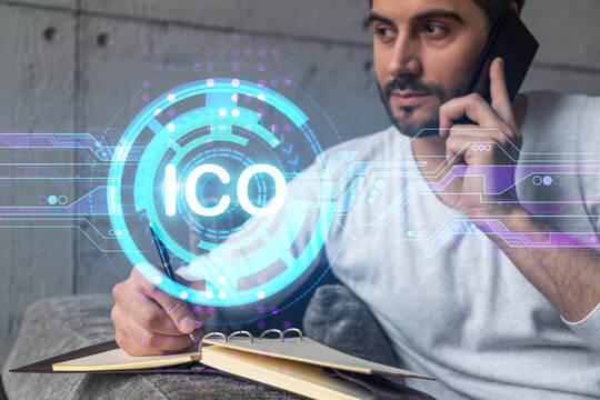 Handsome analyst in casual long sleeve talking phone, taking notes at office workplace try to analyze ICO project. Double exposure. Initial coin offering hologram. - Powered by Adobe