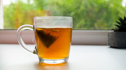 Glass mug with hot tea on the windowsill. Tea bags. Pyramid tea. Steam comes out of the mug. Fragrant delicious tea party. Family pastime.