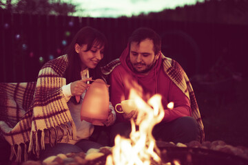 A happy married couple is drinking tea from a teapot outdoors by the fire. Rest in the country with a camper during quarantine. Relaxing near the fire in the nature concept.