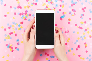 A smartphone in woman hands with perfect manicure. Colourful confetti background. The screen is a...