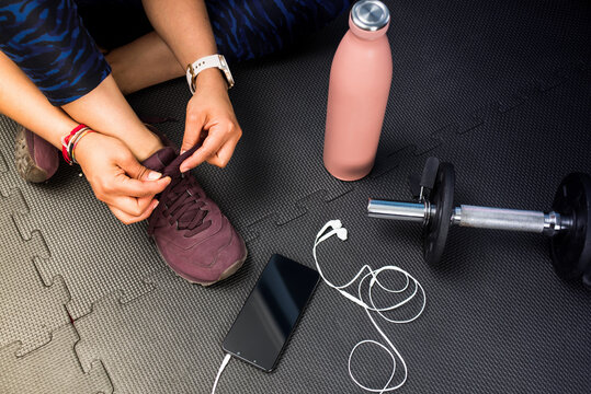 Detail of woman tying footwear to do exercise. Black rubber floor mat and tiles inside a gym. Technology sport with smart phone and earphones.