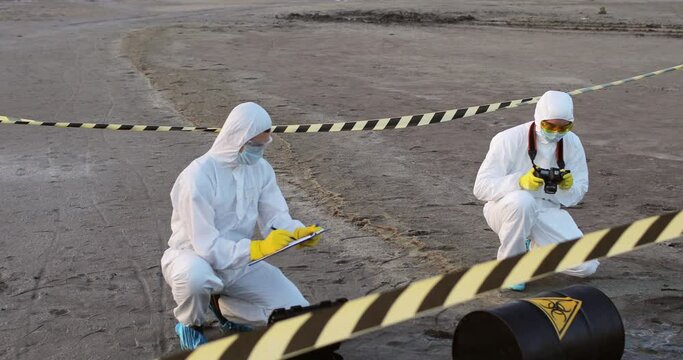 Two scientists in protective suits work in a fenced-in area containing a black barrel with a biohazard sign.