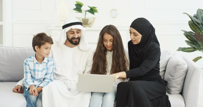 Muslim happy small children sitting with parents on couch and watching video on laptop computer. Mother father with little kids, daughter and son talking, using device. Arabian family on lockdown.