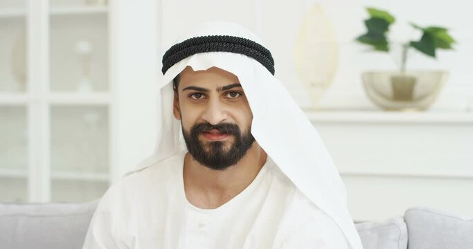 Close up of handsome young Arab man in kandura sitting on couch in living room and looking to camera with smile. Male muslim in dishdasha on sofa at home. Portrait of Arabic millioner from UAE.