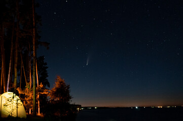 Tourist hikers tent near the lake at night with stars and Neowise comet with light tail in dark...