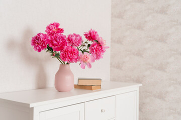 A large bouquet of peonies in a pink vase in a bright room on a white chest, books are nearby. fresh flowers at home, greenhouse, minimalist, cozy loft interior