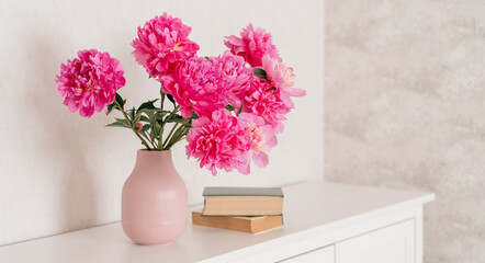 A large bouquet of peonies in a pink vase in a bright room on a white chest, books are nearby. fresh flowers at home, greenhouse, minimalist, cozy loft interior