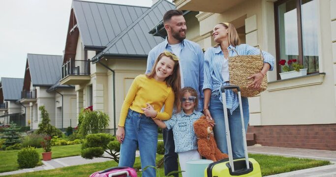 Happy family with things ready to move to another country standing with suitcases against the background of their old sold house and smiling looking at camera. Family preparing for trip.