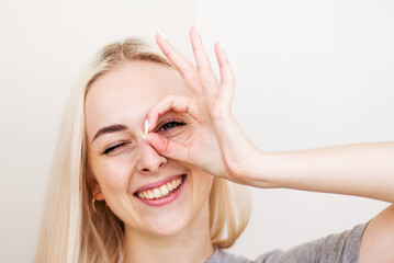 Close up portrait of beautiful joyful blonde female smiling, demonstrating white teeth, looking at the camera through fingers in okay gesture. Face expressions, emotions, and body language