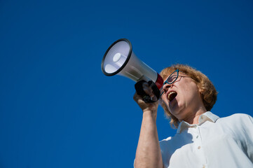 An elderly woman in glasses shouts into a megaphone outdoors. An annoyed pensioner makes her demands with the help of a device amplifying sound against a blue background. Loudspeaker in female hands.