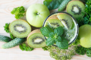 Fototapeta na wymiar Green smoothie in a glass, decorated with mint and kiwi, with apples, kiwi, cucumbers and parsley in background, selective focus, top view