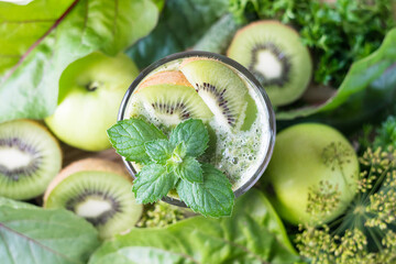 Fototapeta na wymiar Green smoothie in a glass, decorated with mint and kiwi, with apples, kiwi, cucumbers and parsley in background, selective focus, top view