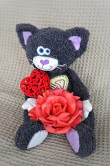 toy plush cat sits on a light background and holds a red rose and a heart. Template for postcard to the day of lovers. High quality photo