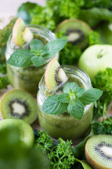 Fototapeta na wymiar Green smoothie in two glass jars with mint and kiwi, with apples, kiwi, cucumbers and parsley in background, selective focus