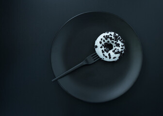 B&W donut on a black plate with a black plastic fork. Monochrome picture of an unhealthy sweet treat.