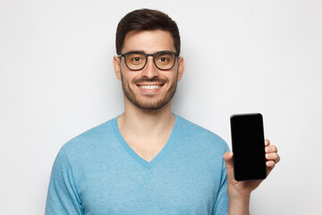 Young man in blue t-shirt and glasses holding blank screen phone in vertical position, isolated on gray background