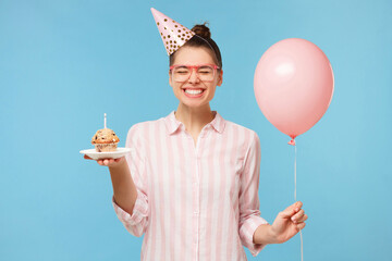 Young happy birthday girl in hat, holding pink balloon in one hand and holiday cupcake in another,...