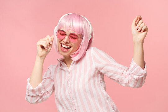 Young Happy Laughing Girl With Pink Hair And Glasses, Listening To Music In Wireless Headphones And Dancing, Isolated On Studio Background