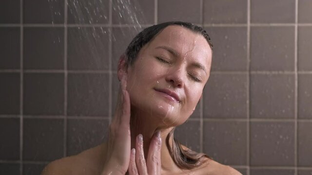 Attractive brunette middle-aged woman takes a shower. Woman washes her hair. Naked body. Grey tile on the walls. Taking care of skin, routine home treatments.