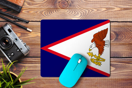 American Samoa flag on wooden background with blue wireless mouse on a mouse pad, top view. Digital media concept.