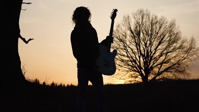 man plays electric guitar and sings a lyric song in a field near the tree at sunset. silhouette