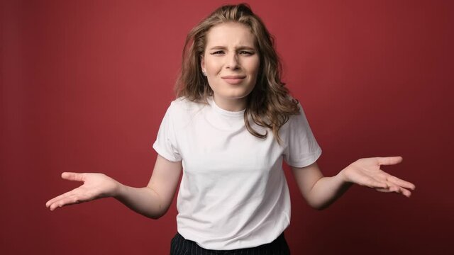 What wrong?! Portrait of confused beautiful girl with curly hair, spreads hands sideways, smirks face, feels doubt while makes choice, dressed in casual t shirt. Isolated on red background.