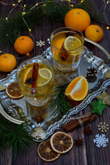 Fototapeta na wymiar Glass cups with aromatic citrus tea, oranges, lemons, cinnamon sticks, decorative snowflakes, Christmas decorations, coniferous branches on a dark wooden background