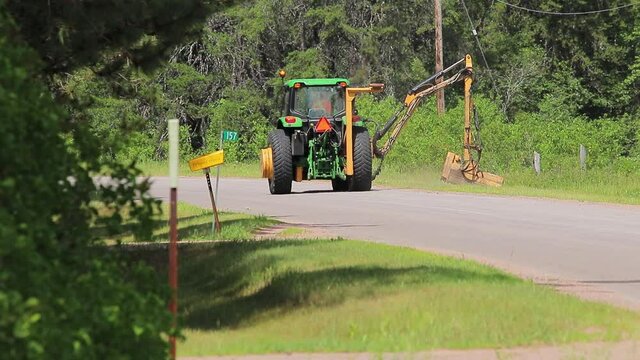Killaloe, Ontario, Canada / July, 23, 2020 :  John Deere tractor cutting brush in ditch of road.  drives around corner out of sight