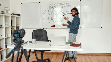 Javascript lesson. Young bearded male teacher wearing glasses pointing at whiteboard and teaching Javascript, giving lesson online. Recording video blog