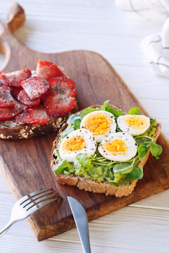 Hard boiled egg on avocado toast with green leaves and toast with fresh strawberries and cream cheese on wooden board, healthy breakfast or lunsh