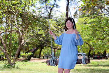 Happy smiling woman in blue dress swinging on a wooden swing in the tropical park. Summer holidays, concept of freedom, happiness and carelessness