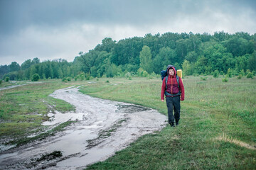 Guy tourist hiker walks along the road with a backpack in a red jacket in the rain