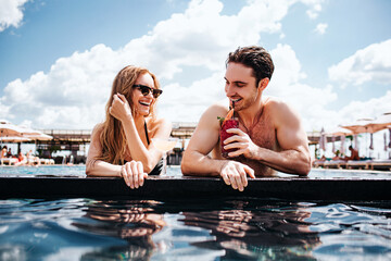 Young hot couple resting at swimpool. Low view of man and woman enjoy rest in resort spa hotel. Look at each other and smile. Drinking cocktails. Summertime vacation.