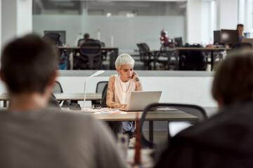 Working on a new project. Focused young and beautiful blonde tattooed businesswoman looking using laptop and thinking about something while sitting at her working place in modern office